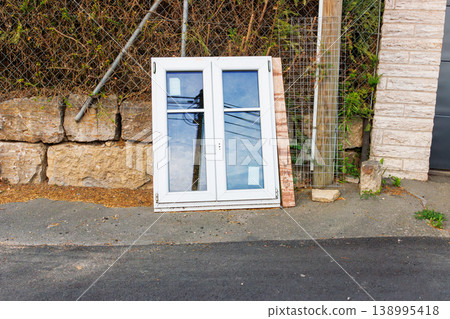 White plastic window frame with glass panes leans against a stone wall beside a road. Discarded building material and renovation debris. White plastic window frame with glass panes leans against a stone wall beside a road. Discarded building material and renovation debris. 138995418