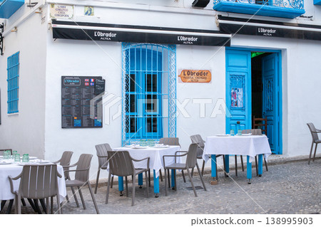 Charming L'Obrador Pizzeria Creperie outdoor dining tables set on a picturesque street in Altea Spain showcasing vibrant blue windows and doors for pleasant travel and tourism family holidays and 138995903