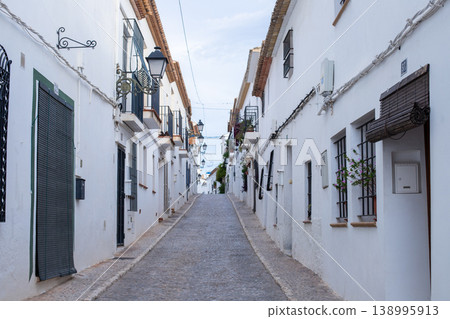 Charming cobblestone street winding uphill through Altea old town with traditional white houses and decorative streetlights under a bright sky, perfect for a Spanish holiday exploration Charming cobblestone street winding uphill through Altea old town with traditional white houses and decorative streetlights under a bright sky, perfect for a Spanish holiday exploration 138995913