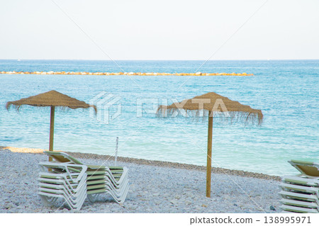 Empty straw umbrellas on the peaceful pebble beach of Altea, Spain, ready for relaxing family holidays and memorable summer vacations by the clear blue sea 138995971