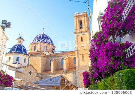 Sunny Altea old town street scene with the famous blue tiled church dome and bell tower framed by abundant purple bougainvillea, perfect for a Spanish holiday 138996008