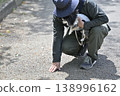 A man walks his dog, a Chihuahua, and checks the ground temperature with his hand. 138996162