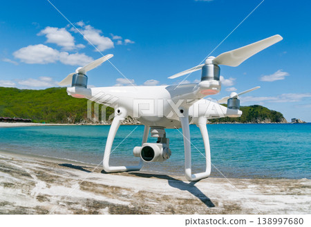 Modern white camera drone positioned on coastal surface overlooking beautiful blue ocean waters and green hills on a sunny day 138997680