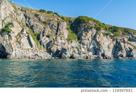 Rugged stone cliffs rise sharply from the deep blue ocean water under a clear, bright summer sky 138997681