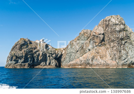 Coastal cliffs featuring a remote white building on its peak, towering over deep blue sea on a bright day 138997686