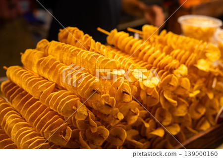 Close-up of crispy, golden spiral-cut tornado potatoes on wooden skewers at a bustling local street food night market. 139000160