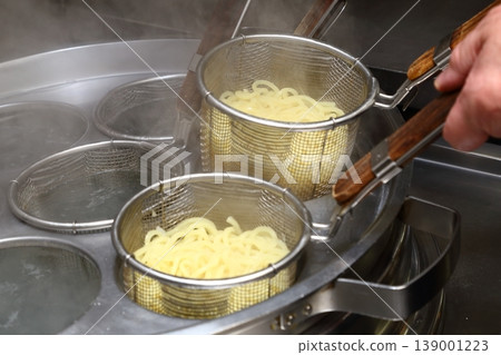 A cooking scene at a ramen shop, where noodles are being lifted (before draining the water from the noodle boiling machine). 139001223