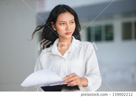 Confident young businesswoman holding a report clipboard in a modern office setting. Professional female manager looking ahead with focus, representing leadership, planning, and corporate success. 139002212