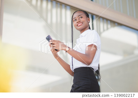 Low angle portrait of young Black business woman holding smartphone and looking away outdoors. Mobile connection, career confidence, online communication, remote work and modern lifestyle concept. Low angle portrait of young Black business woman holding smartphone and looking away outdoors. Mobile connection, career confidence, online communication, remote work and modern lifestyle concept. 139002217