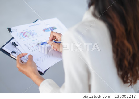 Over-the-shoulder view of a businesswoman pointing to line charts on a report clipboard. Professional female analyst reviewing performance data, financial trends, and business results. Over-the-shoulder view of a businesswoman pointing to line charts on a report clipboard. Professional female analyst reviewing performance data, financial trends, and business results. 139002218
