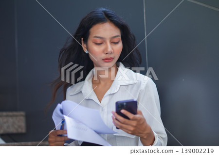 Professional businesswoman looking at smartphone while holding documents in an office stairwell. Female employee checking messages, reviewing paperwork, and coordinating business communication. Professional businesswoman looking at smartphone while holding documents in an office stairwell. Female employee checking messages, reviewing paperwork, and coordinating business communication. 139002219