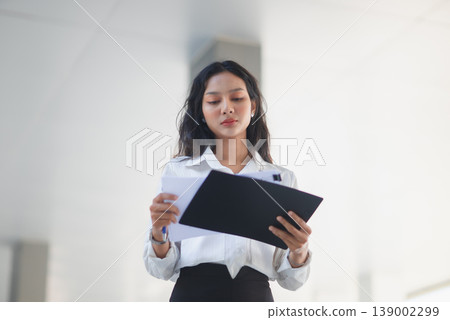 Young professional woman holding a clipboard and documents against a clean white background. Corporate employee reviewing reports, paperwork, and financial data in a minimalist office. Young professional woman holding a clipboard and documents against a clean white background. Corporate employee reviewing reports, paperwork, and financial data in a minimalist office. 139002299