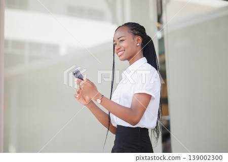 Side view of young Black business woman using smartphone outside an office building. Mobile communication, urban lifestyle, remote work, online messaging and modern professional concept. Side view of young Black business woman using smartphone outside an office building. Mobile communication, urban lifestyle, remote work, online messaging and modern professional concept. 139002300