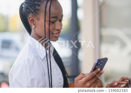 Close portrait of young Black woman using smartphone at an outdoor cafe, focused on the screen. Mobile messaging, online browsing, digital communication and modern lifestyle concept. Close portrait of young Black woman using smartphone at an outdoor cafe, focused on the screen. Mobile messaging, online browsing, digital communication and modern lifestyle concept. 139002385