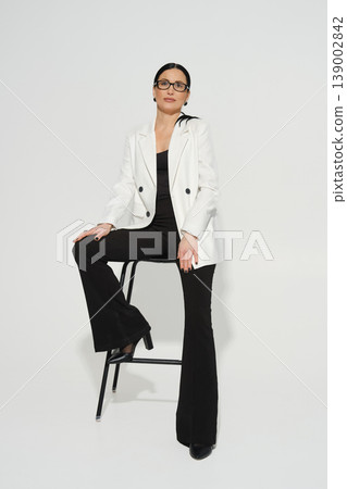Woman sitting on stool wearing a stylish outfit in a plain background during a fashion shoot Woman sitting on stool wearing a stylish outfit in a plain background during a fashion shoot 139002842