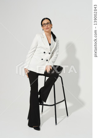 Woman sits on stool wearing a white jacket and black pants in a studio setting during daytime Woman sits on stool wearing a white jacket and black pants in a studio setting during daytime 139002843