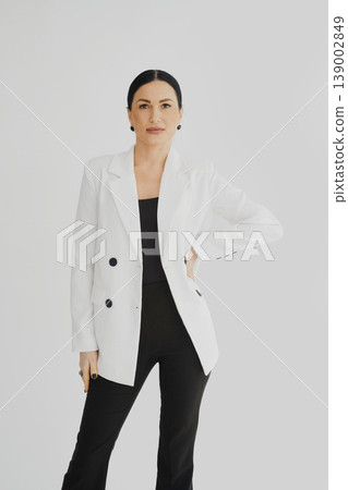 Woman stands confidently in a white blazer and black outfit in a bright room Woman stands confidently in a white blazer and black outfit in a bright room 139002849