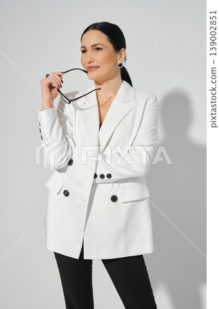 Woman poses in a white blazer while holding sunglasses in a studio setting during daylight Woman poses in a white blazer while holding sunglasses in a studio setting during daylight 139002851