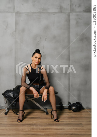Woman sitting on a weight bench in fitness studio with weights nearby in the evening Woman sitting on a weight bench in fitness studio with weights nearby in the evening 139002865