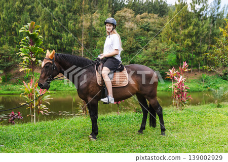 Young woman sitting on horse near tropical lake during riding lesson, beginner equestrian enjoying outdoor activity in countryside Young woman sitting on horse near tropical lake during riding lesson, beginner equestrian enjoying outdoor activity in countryside 139002929