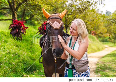 Young woman gently stroking horse in tropical garden, emotional connection with animal during outdoor activity Young woman gently stroking horse in tropical garden, emotional connection with animal during outdoor activity 139002935