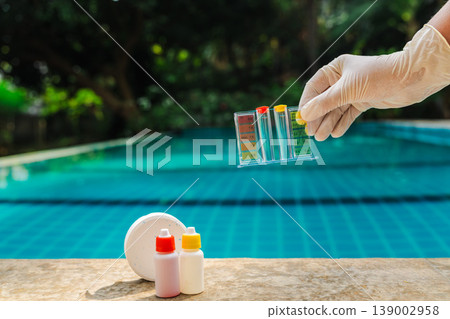 Gloved hand holding pool water test kit above swimming pool with pH chart, chlorine tablet, reagent bottles and clear blue water, pool maintenance scene 139002958