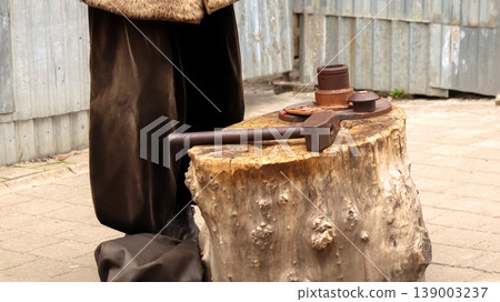 Close-up of a large old hammer lying on a tree stump anvil. Historical reenactment of traditional coin minting or blacksmithing outdoors. Craftsmans tools on a wooden block Close-up of a large old hammer lying on a tree stump anvil. Historical reenactment of traditional coin minting or blacksmithing outdoors. Craftsmans tools on a wooden block 139003237