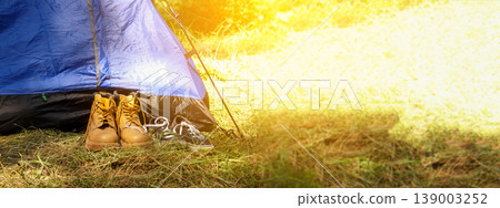 Tourist boots and sneakers standing near a blue tent in a forest. Concept of camping, hiking, and outdoor summer adventures. Panoramic shot with bright sunlight and copy space 139003252