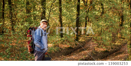 Man with a large backpack and a bucket walking on a dirt road through an autumn forest. Side view of a hiker exploring nature during golden hour. Wide panoramic shot with copy space 139003253