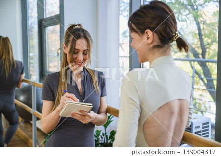 Female trainer taking notes while talking to client in fitness studio. Female trainer taking notes while talking to client in fitness studio. 139004312