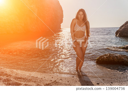 Woman sunset beach standing in shallow ocean waves during golden hour wearing white lingerie on a scenic rocky coastline Woman sunset beach standing in shallow ocean waves during golden hour wearing white lingerie on a scenic rocky coastline 139004736
