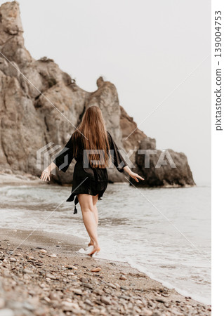 Woman beach ocean walking along the pebbled shoreline with outstretched arms towards a rocky coastline under a misty sky Woman beach ocean walking along the pebbled shoreline with outstretched arms towards a rocky coastline under a misty sky 139004753