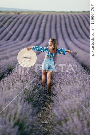 Lavender woman field walking through purple flower rows holding a large straw hat in a blooming countryside landscape Lavender woman field walking through purple flower rows holding a large straw hat in a blooming countryside landscape 139004767