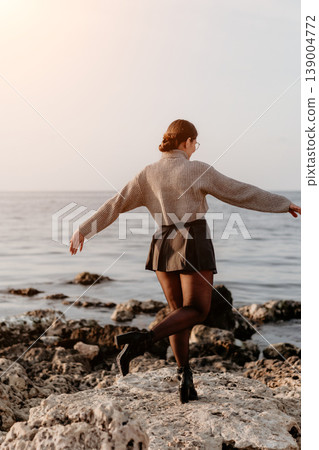 Woman coast sunset balances on rocks with arms outstretched wearing a sweater and skirt in the golden evening light Woman coast sunset balances on rocks with arms outstretched wearing a sweater and skirt in the golden evening light 139004772