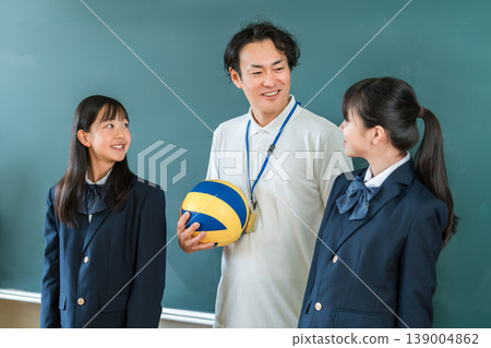 A male teacher holding a volleyball and a female student wearing a blazer in front of a blackboard in a classroom 139004862