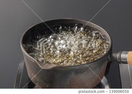 A scene of water boiling vigorously in an iron pot on a gas stove against a black background, with steam rising. An image representing cooking, heating, and boiling. A scene of water boiling vigorously in an iron pot on a gas stove against a black background, with steam rising. An image representing cooking, heating, and boiling. 139006050