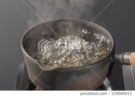 A scene of water boiling vigorously in an iron pot on a gas stove against a black background, with steam rising. An image representing cooking, heating, and boiling. 139006051