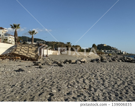 Sunny beach scene with palm trees, pebbled sand, and distant buildings on a clear day background Sunny beach scene with palm trees, pebbled sand, and distant buildings on a clear day background 139006835
