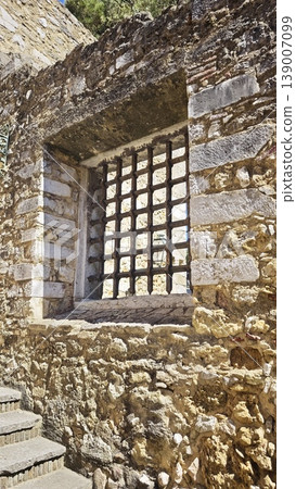 Stone wall displays a barred window with a metal grid, highlighting rustic architecture and historical features in a Mediterranean environment 139007099