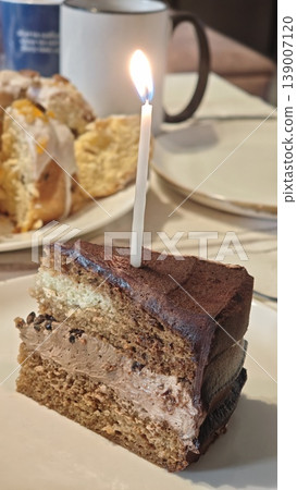 Chocolate cake slice with a lit candle on a white plate, accompanied by scones and a coffee mug in a cozy indoor setting 139007120
