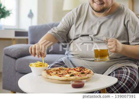 Overweight man holding beer glass mug, eating fries beside pizza at home 139007391