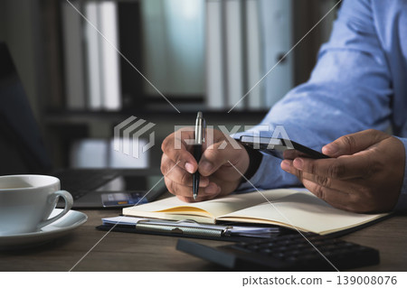 Hand holding smartphone while writing notes in notebook beside laptop, card, calculator, and coffee cup on desk, showing content planning, digital research, copywriting workflow, and remote 139008076