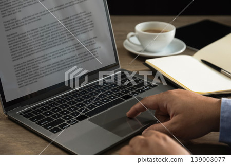 Hand using laptop trackpad with text document beside notebook, tablet, and coffee cup on desk, representing content writing, copy editing, digital publishing, article review, and remote workflow 139008077