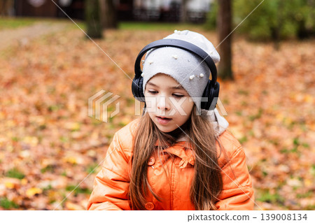 Young girl in orange coat wearing headphones in an autumn park among fallen leaves Young girl in orange coat wearing headphones in an autumn park among fallen leaves 139008134