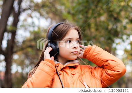 Young girl wearing headphones and an orange jacket listening to music outdoors in an autumn park Young girl wearing headphones and an orange jacket listening to music outdoors in an autumn park 139008151