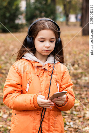Girl with headphones using smartphone in orange coat outdoors in autumn park Girl with headphones using smartphone in orange coat outdoors in autumn park 139008152