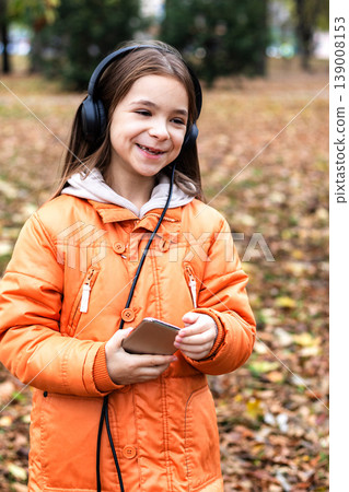 Girl in orange coat listening to music on headphones and smiling in an autumn park Girl in orange coat listening to music on headphones and smiling in an autumn park 139008153