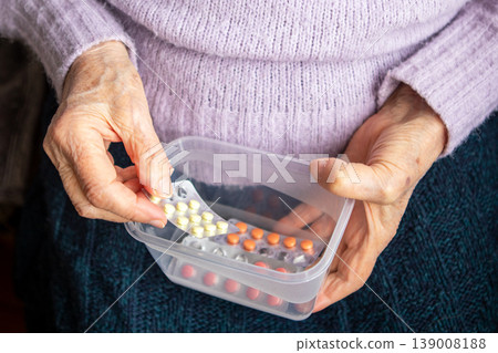 Elderly woman organizing daily medication pills in plastic container with careful hand 139008188