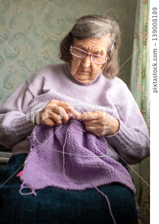 Grandmother knitting a purple sweater at home, focused elderly woman crafting with yarn Grandmother knitting a purple sweater at home, focused elderly woman crafting with yarn 139008189