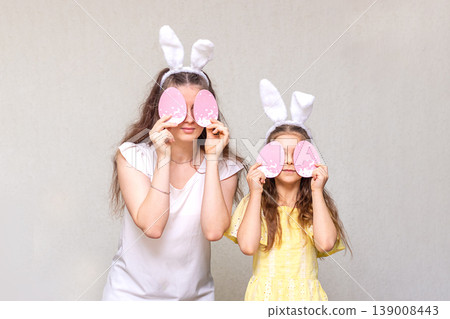 Mother and daughter wearing bunny ears holding pink Easter egg masks over their eyes 139008443